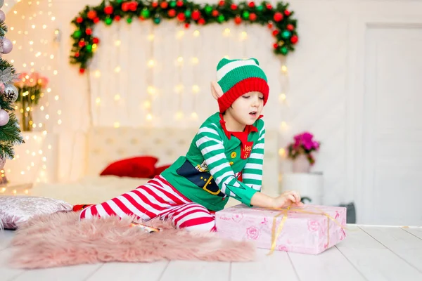 Joyeux garçon habillé comme un gnome assis sur le sol dans son salon pendant la période de Noël. Enfant tient un cadeau de Noël. — Photo