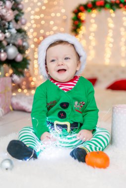 Noel hediyeleri ve oyuncaklarıyla elf gibi giyinmiş bir çocuk. Zarif bir Noel ağacının yanında hediye kutusu ve şekeri olan küçük bir çocuk. Aile kış tatilini kutluyor. Ev dekorasyonu.