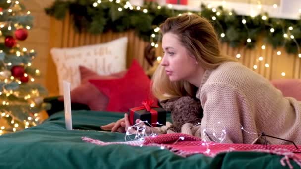 Jovem loira conversando na rede com um laptop, fica na cama perto de uma árvore de Natal decorada com uma caixa de presente em suas mãos. Uma menina escolhe um presente de Ano Novo pela Internet — Vídeo de Stock