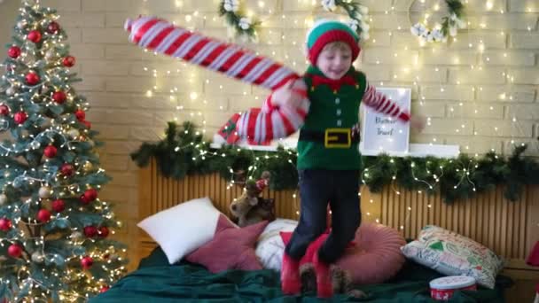 Маленький веселый мальчик в шляпе эльфа прыгает на кровать с надувной тростью в руках — стоковое видео
