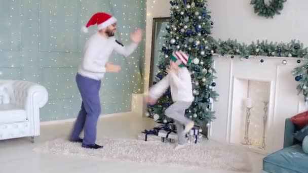 Tata tańczy ze swoim synkiem, ciesząc się Bożym Narodzeniem razem w ozdobnym pokoju w domu z jasno oświetlonymi światłami i choinką. — Wideo stockowe