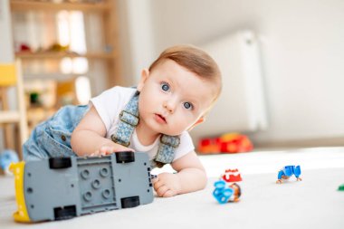 Kameraya bakıp, oyun odasında oyuncak otobüsle oynayan sevimli bir bebek.