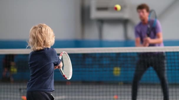 A man coach teaches a little boy to play on an indoor court. A professional tennis instructor throws the ball to hit the racket to the kid, how to return the ball with the racket — Stock Video