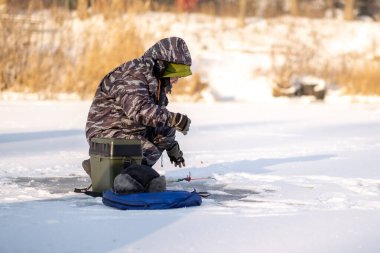 Bir balıkçı bir kutuya oturur ve kışın güneşli havada balık yakalar.