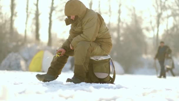 Um pescador senta-se em uma caixa e captura peixes no inverno em tempo ensolarado — Vídeo de Stock