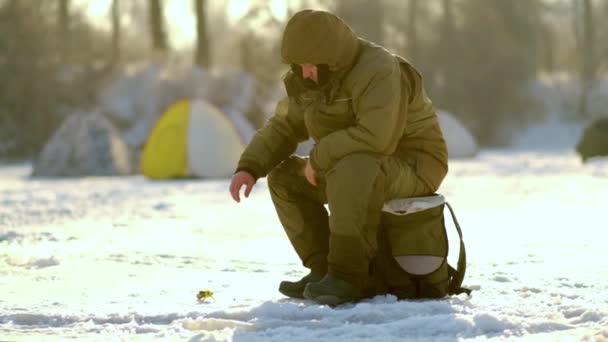 Um pescador senta-se em uma caixa e captura peixes no inverno em tempo ensolarado — Vídeo de Stock