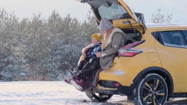 Roztomilá rodinná matka a syn sedí v otevřeném kufru svého žlutého auta a popíjejí teplý nápoj, užívají si krásnou přírodu zasněžené zimy, odpočívají se psem na západě vysočiny bílý teriér, zpomalený pohyb — Stock video