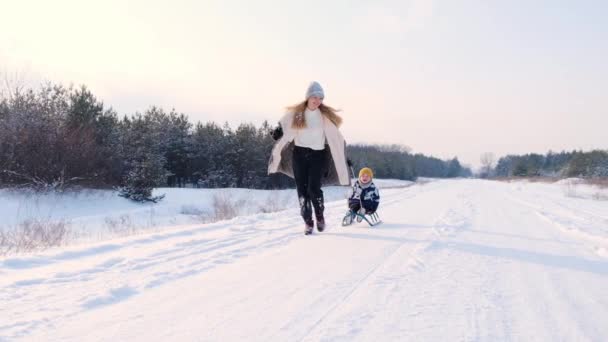 Θετική παιχνιδιάρικη ξανθιά μητέρα τρέχει φέρνοντας χαριτωμένο μικρό γιο της στο έλκηθρο, οικογενειακή ανάπαυση και να διασκεδάσουν περνώντας χρόνο μαζί στο δάσος κατά τη διάρκεια χιονοπτώσεων, χειμερινές δραστηριότητες, αργή κίνηση — Αρχείο Βίντεο