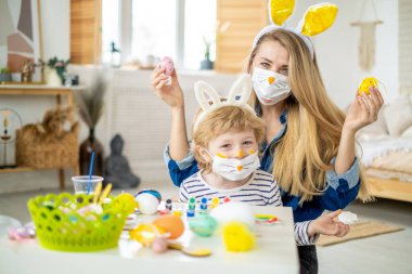 Koronavirüs salgını sırasında maskeli bir aile Paskalya yumurtası avında eğleniyor. Anne ve oğul, koronavirüs salgınını kutlamak için tavşan kulağı ve tavşan kostümü giyiyor. İyi tatiller..