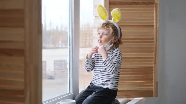 Концепция Пасхи 2021: Ребенок в домашнем карантине играет у окна с ушами кролика на голове в медицинской маске против вирусов во время коронавируса COVID-2019 и вспышки гриппа. — стоковое видео