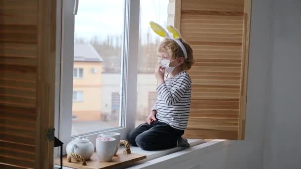 Pasen 2021 concept: Een kind in eigen huis quarantaine speelt bij het raam met de oren van een konijn op zijn hoofd met een medisch masker tegen virussen tijdens het coronavirus COVID-2019 en de griepuitbraak. — Stockvideo