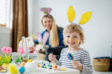 Saçbantlı, tavşan kulaklı güzel anne ve oğul. Yumurtaları fırça ve parlak boyalarla süsler. Paskalyayı kutlamaya hazırlan, evde eğlen.