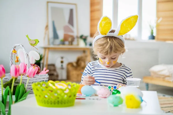 Tavşan kulaklı ve yüzlü şirin çocuk Paskalya bayramı için boya ve fırçayla yumurta boyama, yumurta süsleme, yaratıcılık ve hayal gücü geliştirme, mutlu bir şekilde evde zaman geçirme.