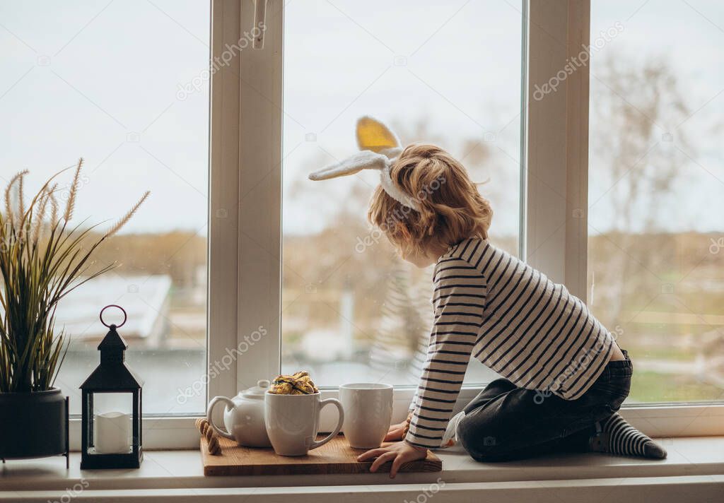 Cute little boy toddler with bunny ears headband sitting on big cozy and pretty decorated windowsill, l oking at window, enjoying view outdoor, feeling comfort with reliable window systems