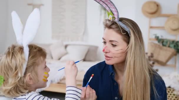 Hermoso hijo feliz y madre en diademas con orejas de conejo decorar los huevos con un pincel y pinturas brillantes, prepararse para celebrar la Pascua, divertirse en casa — Vídeos de Stock