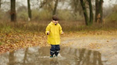 Parkta yağmurlu bir sonbahar gününde su birikintilerinde zıplayan sarı yağmurluk giymiş mutlu çocuk..