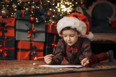 Noel Baba şapkası takan ve papyonlu ekose gömlek giyen bir çocuk Noel Baba 'ya parlak Noel ağacı ışıklarının altına bir mektup yazıyor. Saf tatil büyüsü. 