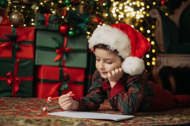 Noel Baba şapkası takan ve papyonlu ekose gömlek giyen bir çocuk Noel Baba 'ya parlak Noel ağacı ışıklarının altına bir mektup yazıyor. Saf tatil büyüsü.
