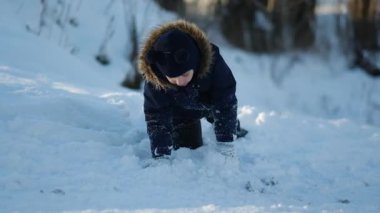 Koyu mavi ceketli, kürklü ve ponpon şapkalı küçük bir çocuk ormanda mutlu bir şekilde karla oynuyor. Eldivenlerini çırpıyor, karı dağıtıyor, gülümsüyor ve zevkle gözlerini kapıyor. Güneşli bir kış günü..