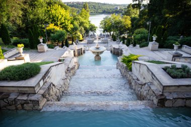 Moldova 'da huzurlu bir parkta terastan akan taştan bir çeşme. Yeşillik ve sakin bir göle bakan bu manzara doğal güzelliği, yumuşak ışığı ve sakin atmosferi yakalar. Seyahat için ideal