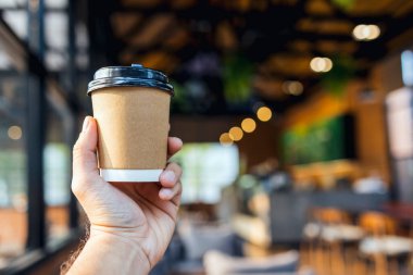 Yakın plan, kafeteryada kağıt kahve bardağını tutarken arka planda hafif bir odaklanma ve ışık var.
