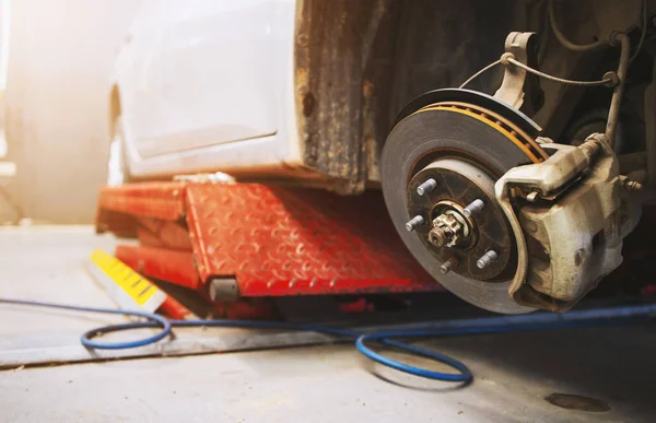 closeup car on a lift in a car service with a wheel removed. the technician is checking disk brake and brake caliper with soft-focus and over light in the background
