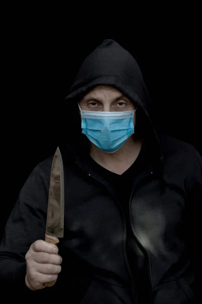 Man hidden in hood wearing  medical mask holding a knife and looking at the camera isolated on black background. Robbery, violence and economic crisis  concept during the covid-19 pandemic. Black and white photography