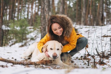 Sarı ceketli gülümseyen genç bir kadın ve büyük beyaz köpeği olan Labrador kış ormanında yürüyor.