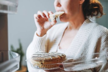 Çekici, dost canlısı, orta yaşlı, rahat hırkalı esmer kadın mutfaktaki buzdolabından pasta yiyor.