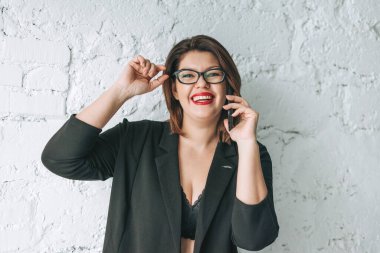 Güzel gülümseyen genç bayan iş kadını gözlüklü ve siyah ceketli, cep telefonu beyaz duvara dayalı.