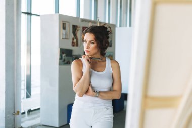 Modern sanat galerisindeki sehpanın önünde beyaz elbiseli, boya fırçalı çekici genç bir kadın ressam düşünüyorum.