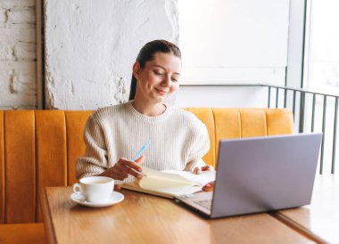 Çekici, genç, esmer, gülümseyen, bir fincan çayla serbest çalışan, kafede laptopunu kullanan bir kadın öğrenci.