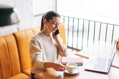 Çekici, genç, esmer, gülümseyen, bir fincan çayla serbest çalışan, kafede çalışan, cep telefonu ve dizüstü bilgisayar kullanan bir kadın öğrenci.
