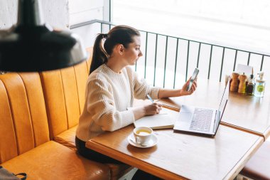 Çekici, genç, esmer, gülümseyen, bir fincan çayla serbest çalışan, kafede çalışan, cep telefonu ve dizüstü bilgisayar kullanan bir kadın öğrenci.