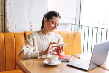 Çekici, genç, esmer, gülümseyen, bir fincan çayla serbest çalışan, kafede çalışan, cep telefonu ve dizüstü bilgisayar kullanan bir kadın öğrenci.