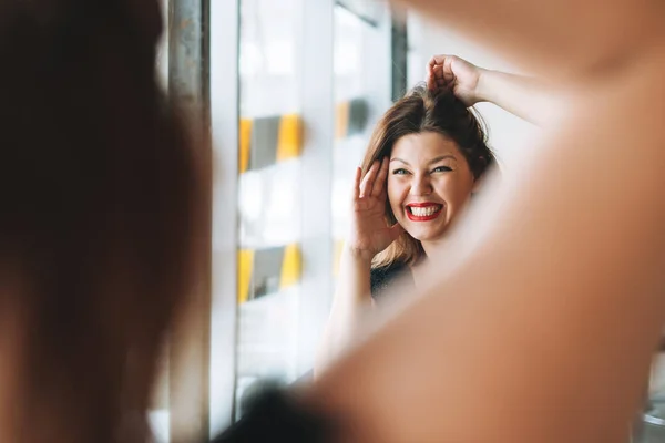 Beautiful smiling young woman plus size body positive in black evening dress make hair style near mirror at beauty studio