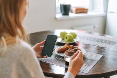 Güzel sarışın kadın kahvaltı yapıyor ve evdeki oturma odasında cep telefonu kullanıyor.