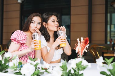 Mutlu gülümseyen, güzel esmer, yaz kıyafetleri içinde ellerinde meyve suyuyla yaz kenti caddesindeki kafede oturan güzel bayan arkadaşlar.