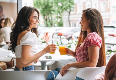 Mutlu gülümseyen, güzel esmer, yaz kıyafetleri içinde ellerinde meyve suyuyla yaz kenti caddesindeki kafede oturan genç bayan arkadaşlar.