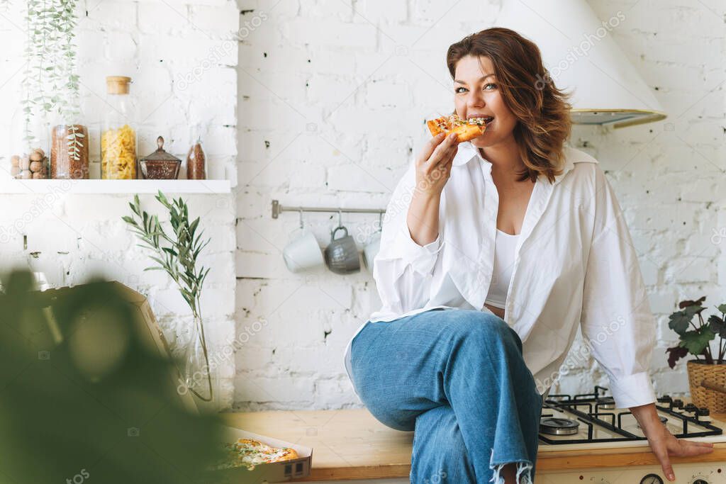 Gorgeous young woman plus size body positive in blue jeans and white shirt eating pizza in home kitchen
