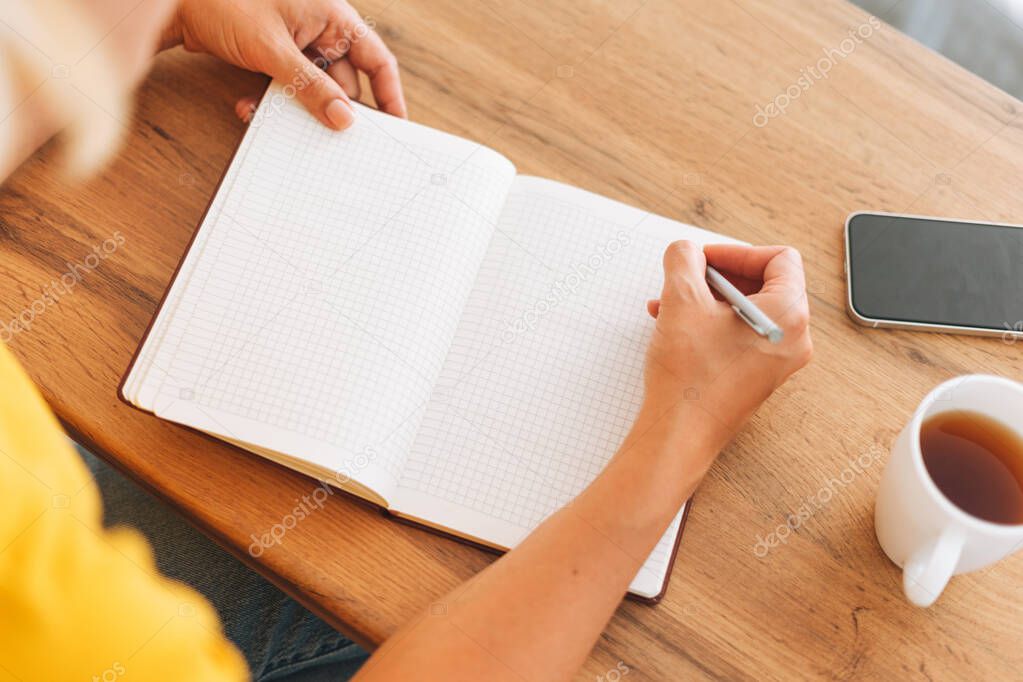 Young blonde woman in yellow t-shirt counts family budget and makes notes in notebook with cup of tea in kitchen at home, top view