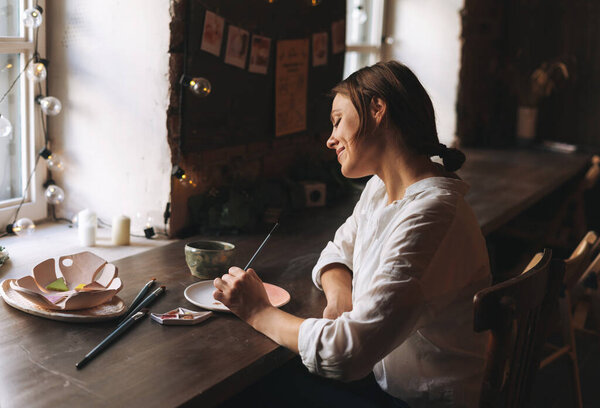 Young attractive woman in white shirt ceramic artist decorating clay plate with tool at table in pottery workshop. Handmade work student, freelance small business concept