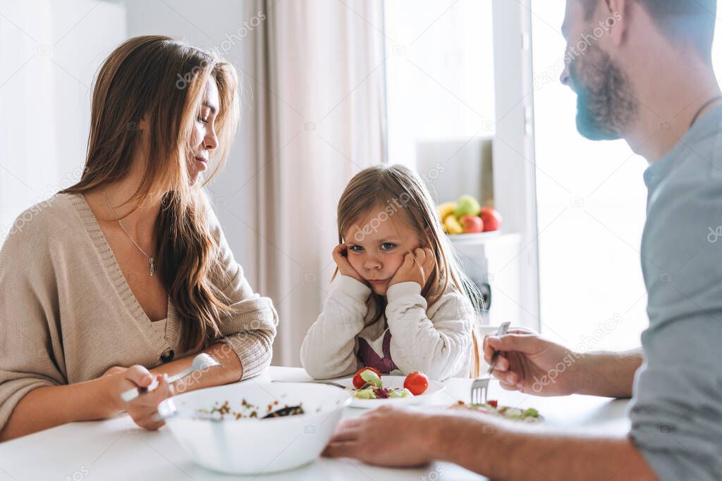 Young family has lunch at table in bright kitchen at home. The daughter does not want to eat and parents are worried