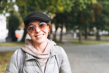 Güneşli bir sonbahar gününde şehir parkında şapkalı ve güneş gözlüklü mutlu, modaya uygun genç bir kadının portresi.
