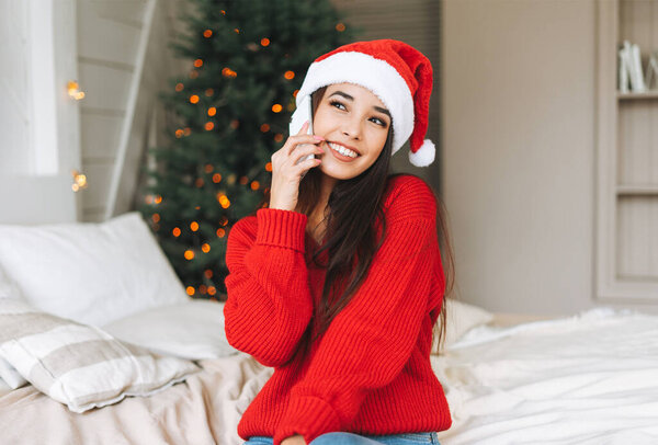 Young beautiful asian woman with dark long hair in cozy red knitted sweater and santa hat using mobile smartphone on the bed in room with Christmas tree
