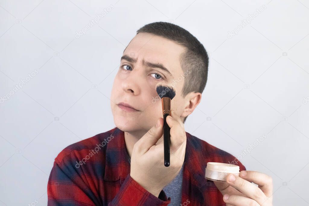 The man looks into the frame and uses powder. Close-up of a guy putting makeup on his face. LGBT community, gay or self-care concept.