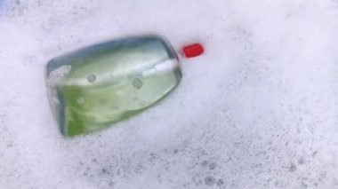 Köpükte bir şişe deterjan yüzüyor. Evdeki kimyasalların tehlikeleri kavramı. Köpükteki sodyum ödüllü maddeler, parabenler, sülfatlar ve diğer kimyasal bileşikler. Sarı kavanozda sıvı sabun.