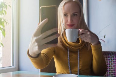 Blogcu kız bir kafede oturur ve akıllı telefondan kapuçino köpüğü atar. Sosyal ağlarda, video bloglarında ve fotoğraf bloglarında çalışıyorum. İnternet içeriği oluşturma ve gıda fotoğrafçılığı kavramı.