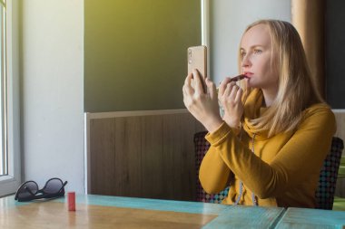 Kız bir kafede dudaklarını boyuyor. Yemekten sonra makyajını yapan tatlı bir kızın yakın çekimi. Kadın çok renkli bir çerçevede bir ayna tutuyor. Kişisel bakım ve makyaj konsepti