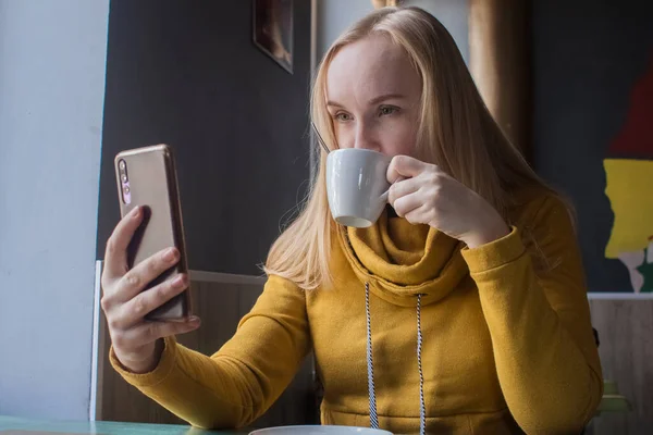 Blogger girl sits in a cafe and shoots a cappuccino foam on the smartphone. Work in social networks, video blogs and photo blogs. Concept for internet content creation and food photography.